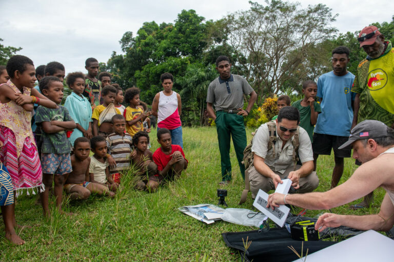 large group of children and a few adults from Papua watch as two crouching white men try to open technical tools in a field.