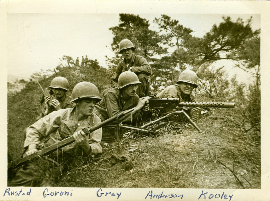 an old photo, discolored yellow showing 5 American soldiers in combat helmets setting up machine guns and rifles on a little hill. One soldier in the background has a large radio held to his ear.