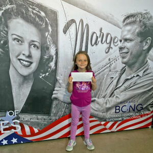 small girl in fuschia shirt and pink pants holding a white envelope standing in front of a black and white panel of a woman on the left side of the girls and on the right is a man in a World War 2 military uniform